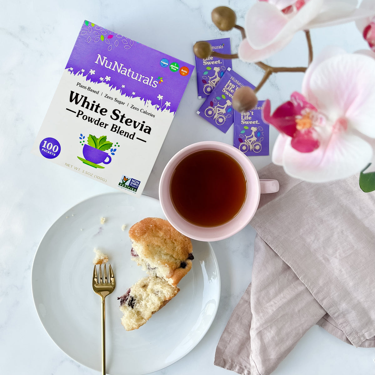 White Stevia Powder Blend Packets box and loose packets beside a cup of tea and a blueberry muffin on a plate
