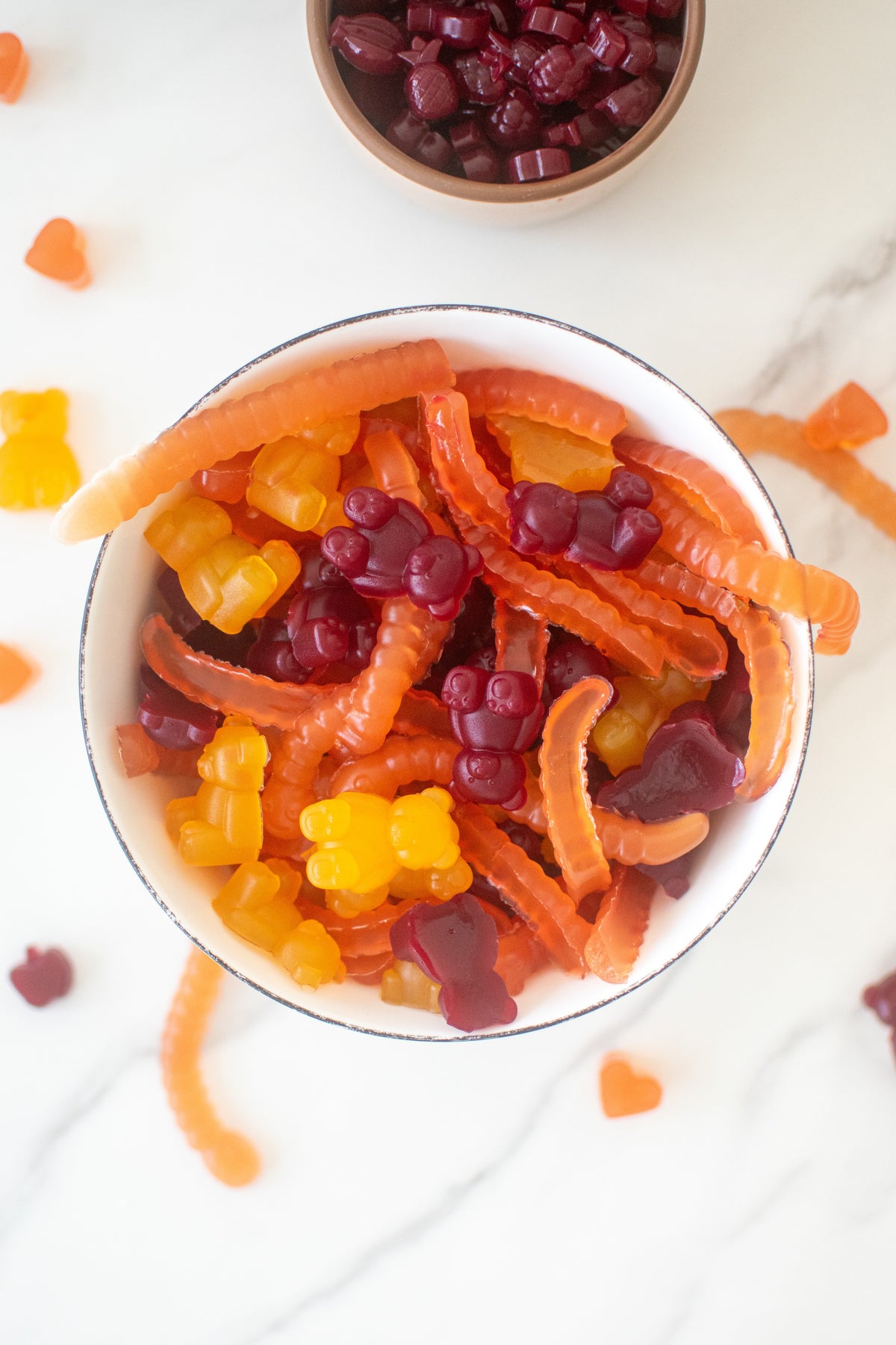 Bowl of orange, yellow, and purple gummy bears and gummy worms on marble, viewed from above